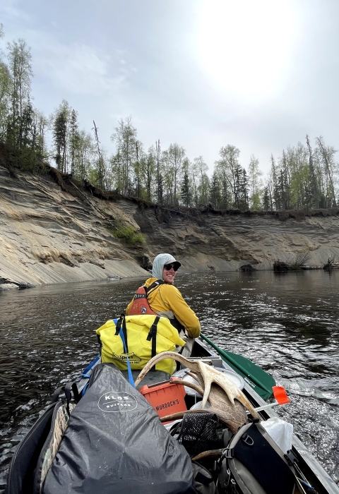 Tim Ericson, sitting at the front of a boat, paddles on the Deshka River. The photo is taken from the back of the boat, and Ericson looks back at the camera over his shoulder. The boat is filled with supplies, including moose antlers. 