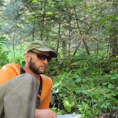 Fish Biologist, Brian Davis, collecting samples in the field. Brian is wearing an orange shirt, tan pants, tan hart and black sunglasses. He is reclining on the ground in a temperate rainforest setting with sampling equipment in front of him.