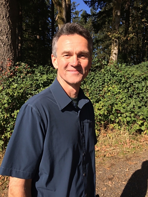 Person stands in sun wearing blue polo with forest in the background.