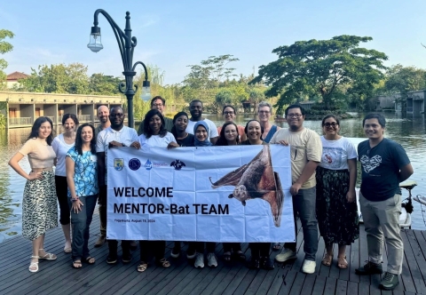 A group of about 18 people stand smiling at the camera holding a large banner that reads "Welcome MENTOR-Bat Team." 