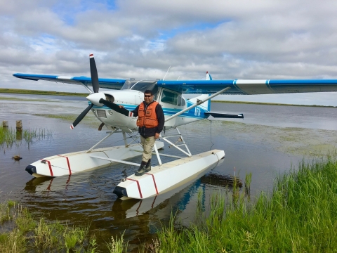 A pilot stands on the floats of a float plane as it floats on a lake shoreline 