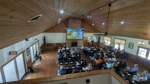 Around 50 people sit in rows of chairs at the center of a large hall. The hall has wooden ceilings with a disco ball hanging from the center. On the far wall a projection screen displays the Tribe's insignia over the mantle of a brick fireplace. 
