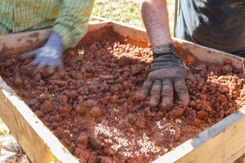 Two gloved hands pushing around chunks of clay in a large sieve