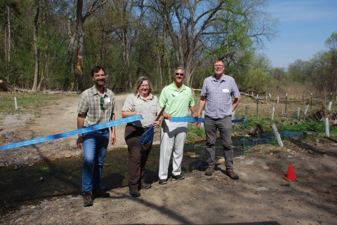  Four people are standing in front of a stream. One of them is holding very large scissors and blue ribbon that has been cut. 