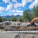 excavator digging near stream with mountains and clouds in background