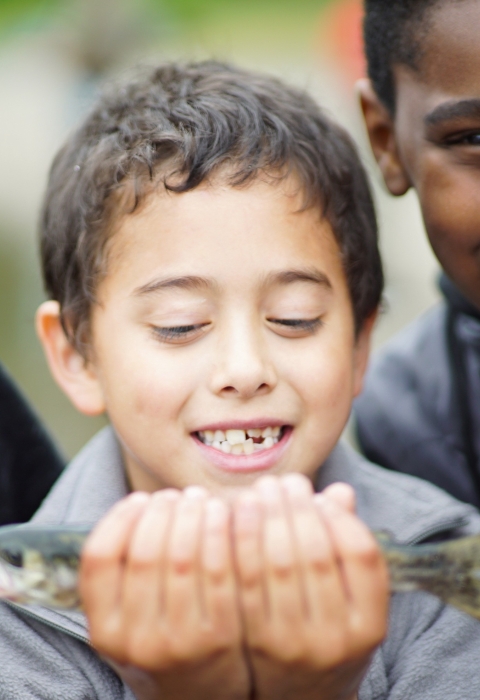 A young child looks at a fish he is holding. A child next to him smiles and points at the camera. 