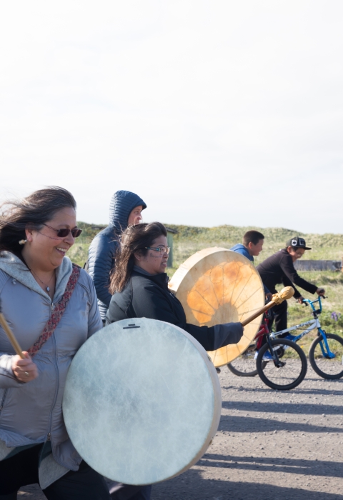 Woman with white skin drum leads group of people walking down a road. Kids on bikes flank her.