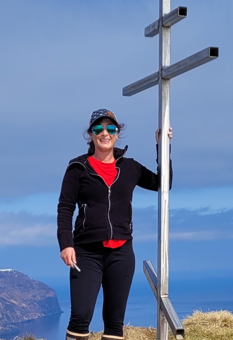 A woman wearing a hat and sunglasses on a high summit overlooking snowcapped mountains and ocean. She stands next to a metal structure.