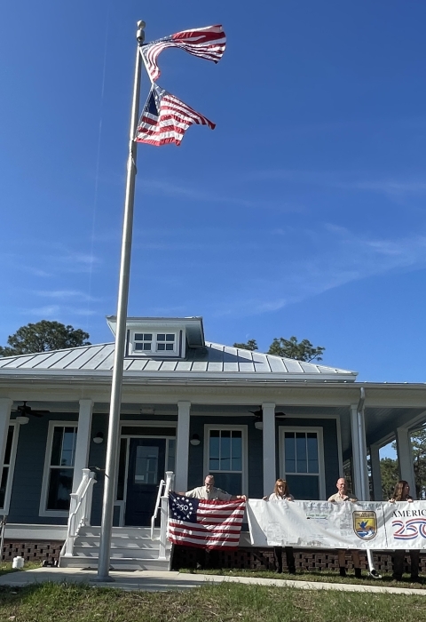 office building with 2 US flags on flagpole in front and people holding banner, Freedom 250 flag