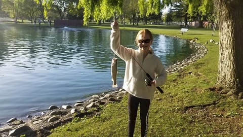 A child stands next to a body of water, holding up a fish and smiling at the camera