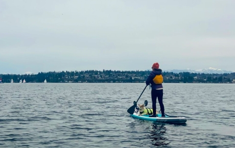 A woman wearing a jacket, pants, and warm hat with a dog on a paddleboard paddling across water.