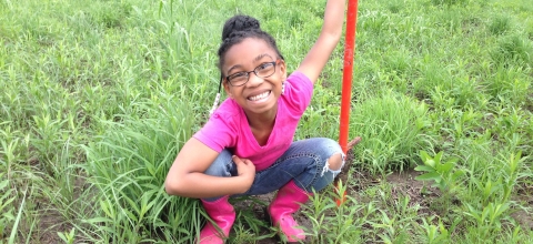 A smiling girl looks up from the grassy area where she is planting milkweed.