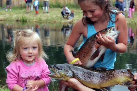 A very young blond-haired girl and an older girl pose with large trout in front of a pond where other children are fishing.