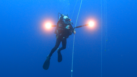 Dr. Peter Auster, weightless in the expanse of this undersea wilderness, pauses to take in the view at 25 m (85 ft) depth, with 1800 m (5940 ft) below him to the summit of Retriever Seamount. He has two large lights to illuminate the darkness and is holding onto a vertical rope / tether.