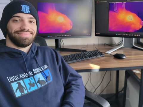 a white man with a beard and mustache wearing a blue stocking knit cap and blue hoodie sits in front of two computer monitors showing a rabbit's head in orange, yellow and purple