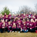 Group of women of varying ages pose for a photo along a roadside while wearing matching maroon sweatshirts made for the trapping workshop. 