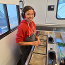 AmeriCorps intern in a fish marking trailer wearing a bib and ear protection. In front of her are four submerged net baskets, two containing juvenile coho salmon.