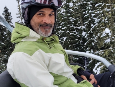 A man smiles while sitting on a ski lift wearing a helmet and green and white jacket. Trees with snow are in the background.