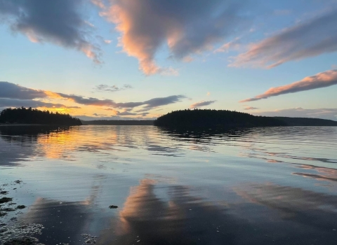Streaks of brightly lit clouds at sunrise reflect on a body of water with distant islands
