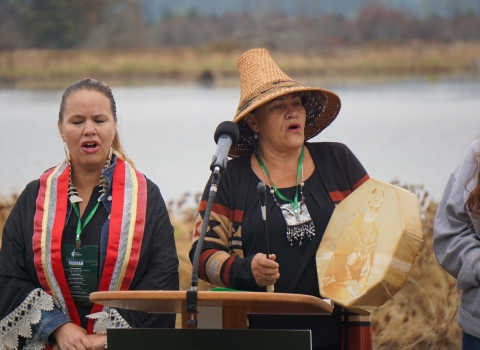 Two native singers, with one wearing a straw hat and holding a drum, behind a podium