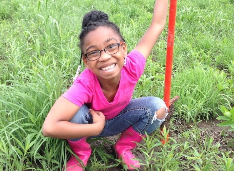 A smiling girl looks up from the grassy area where she is planting milkweed.