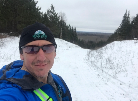 A man in a knit hat, sunglasses and a blue jacket smiles at the top of a snowy trail.