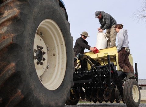 Three people standing on a tractor-pulled seed planter, with a large tractor tire in the foreground.