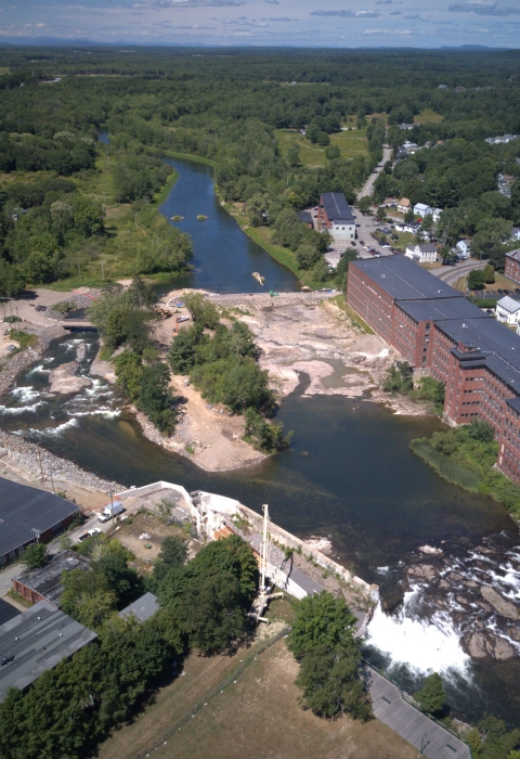 Aerial view of a river flowing through a city. Waterfalls, and a fishway channel are visible. Red brick buildings line the river bank. 