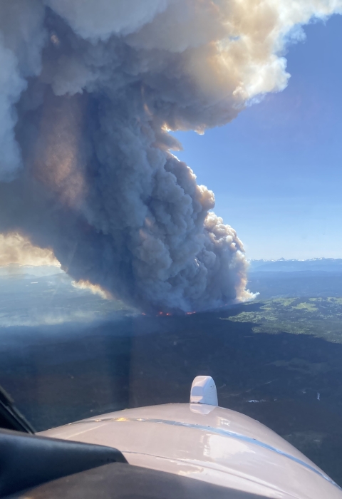 A view of a smoke plume from an airplane. The plume of smoke is billowing into blue sky from a forested landscape.