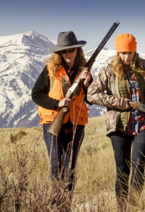 Two women carrying rifles and wearing bright orange gear walk through grass with the Teton Mountains behind them.