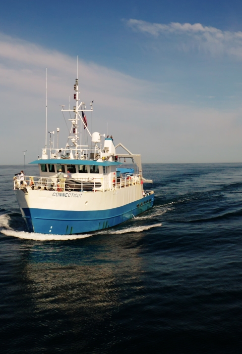 The Research Vessel Connecticut, a 90' steel blue and white ship with a tall mast and various antennas, is traveling through the water with no land in sight. The horizon stretches out far in the distance. The sky is blue, with white clouds.
