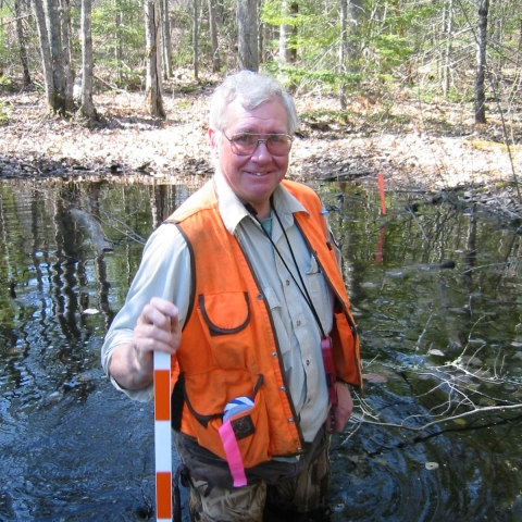 A man holding a yard stick and wearing an orange vest and waders standing in a shallow pond in a forested area