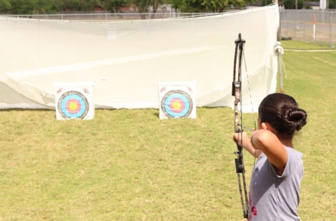 A young woman aims a bow and arrow at a target on a sunny afternoon.
