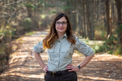 A woman with long brown hair stands on a trail framed by forest. Her hands are on her hips. She wears glasses and a U.S. Fish and Wildlife Service Uniform. 