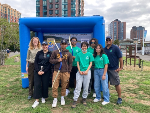 8 people stand in front of a large inflated square object. Some of the people hold archery bows. The group is smiling. It's a cloudy day and there are urban buildings in the background.