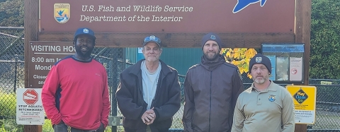 Four men stand smiling in front of a large sign for the Eagle Creek National Fish Hatchery. Sunlight and green grass are behind them.