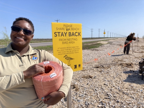 FWS employee smiles at camera and holds a roll of twine while another employee uses the twine to build an exclosure to protect nesting birds