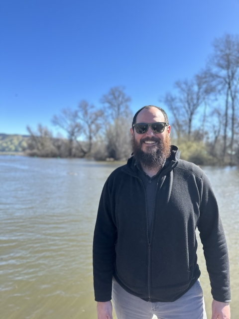 A male biologist with a beard wears sunglasses and a black sweatshirt near a murky lake with blue skies