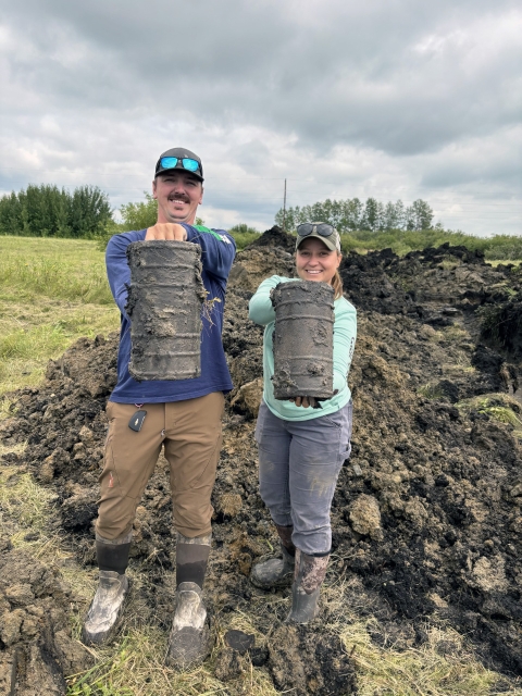 Two adults hold dirt-covered, footlong sections of drain tile with exposed soil in the background. 