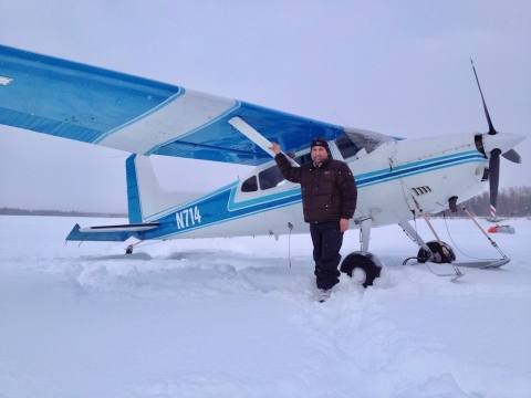 a uniformed officer poses with a single engine plane equipped with skis on its landing gear on a snow covered parking apron 