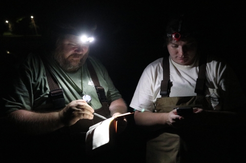 Two people standing in the dark, wearing headlamps. One holds a phone, the other a pen and notebook