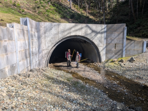 Two people in fishing waders wearing backpacks stand under a large culvert in a streambed