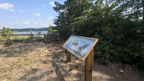 A sign with the title "Dancing Point" depicts Tribal dancers in their ceremonial dress. The wooden sign stands in a clearing overlooking the Rappahannock River. 