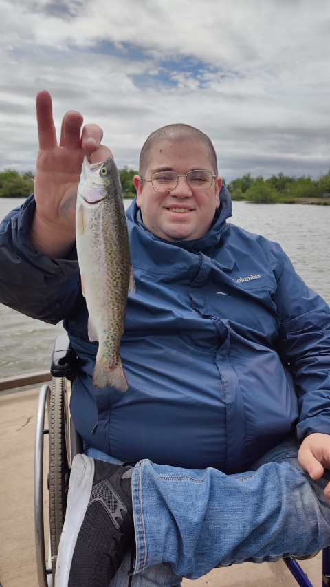 A smiling man in a manual wheelchair holds up a freshly caught trout, wearing a blue jacket and jeans, with cloudy skies and pond in the background.