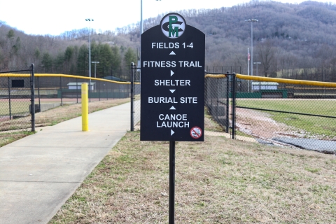 Sign between baseball fields and beside a sidewalk pointing the way to park facilities and a burial site