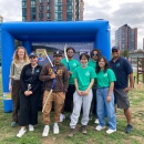 8 people stand in front of a large inflated square object. Some of the people hold archery bows. The group is smiling. It's a cloudy day and there are urban buildings in the background.