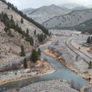 An aerial view over a winding river with a road along one side and mountains with evergreen trees on the other. there is a light dusting of snow on the road and terrain.