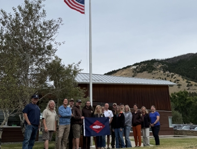 2 US flags fly and at base of flagpole, people pose with fish flag