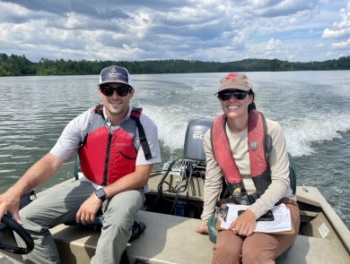 Two people sit on a moving boat, with one person driving while the other holds a clipboard