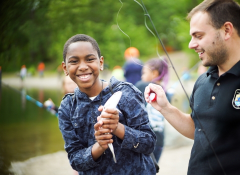 A boy holds a fish in his hands and smiles at the camera. A person next to him holds a fishing rod an bobber.